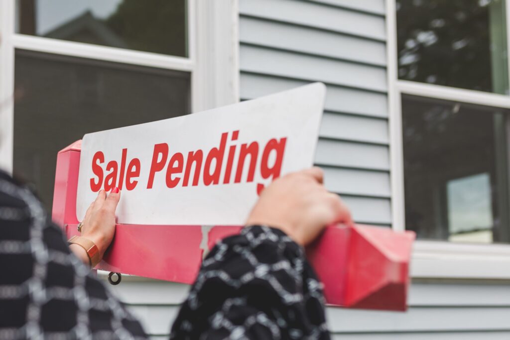 A close-up view from behind a real estate agent's hands as they attach a white sign rider that reads "Sale Pending" in bold red letters onto a pink yard post in front of a house with light grey siding