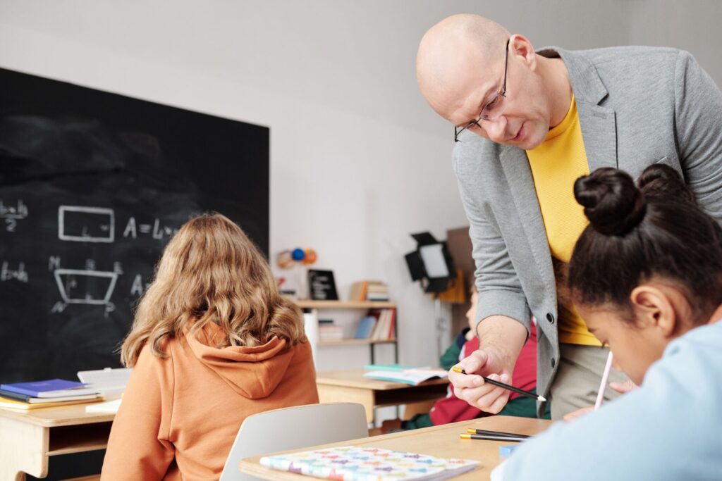 Male teacher in a grey jacket leaning over a student's desk with a pencil to assist during an elementary class