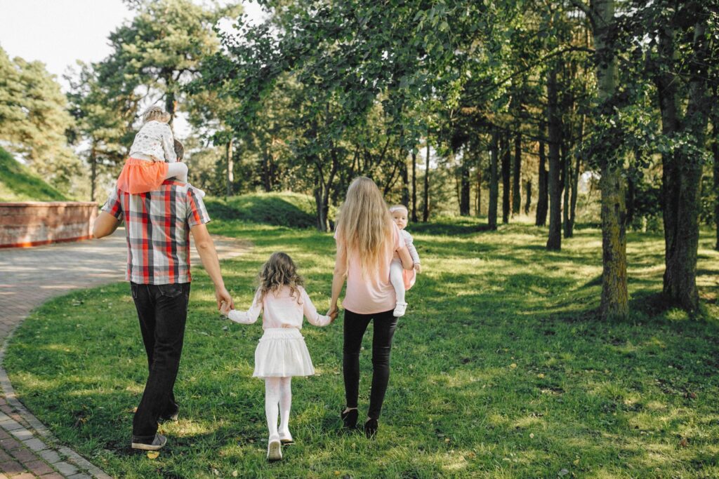 Rear view of a family of five with three young daughters walking along a grassy path through a sunlit park