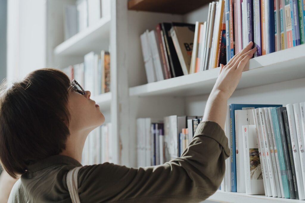 Young woman with glasses reaching for a book on a high shelf in a white library bookcase